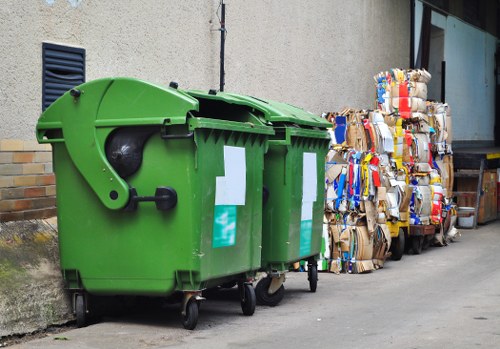 A bright green wheeled commercial waste bin with a closed lid, positioned on a pavement next to a grey concrete wall with a small rectangular ventilation grille. To the right of the bin, there are multiple tightly packed bales of flattened cardboard and paper packaging, stacked against a building with large glass windows. The cardboard bales display various colors and textures, indicating mixed recyclable materials, and are arranged in an orderly pile. The setting appears to be an outdoor area near a commercial or residential property in Shepperton, with a paved ground surface and some visible shadows cast by the objects from natural lighting. The scene reflects typical rubbish and recycling collection preparations handled by a local waste management service, such as House Clearance Shepperton.
