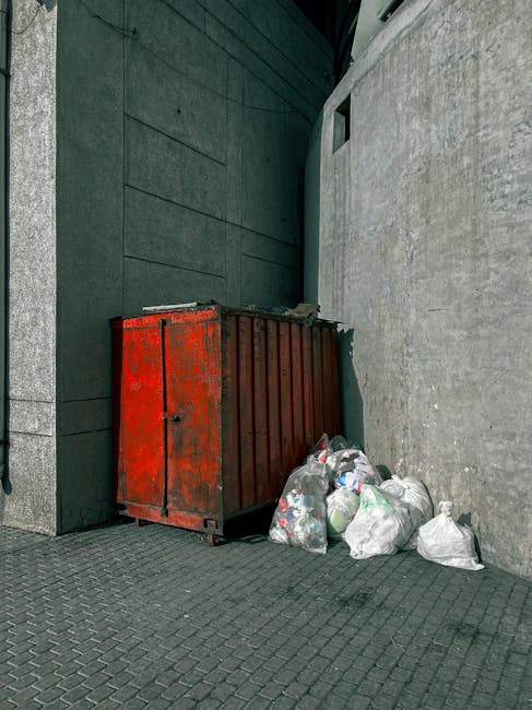 A worker in a high-visibility vest is engaged in rubbish collection at the rear of a large red waste collection vehicle parked on a roadside. The vehicle's rear section is open, revealing mechanical c