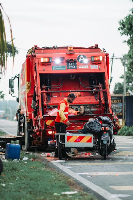 A worker in a yellow vest and dark trousers operates a large red rubbish collection vehicle on a roadside, with the vehicle's rear hatch open, revealing the internal compactor mechanism. The worker is